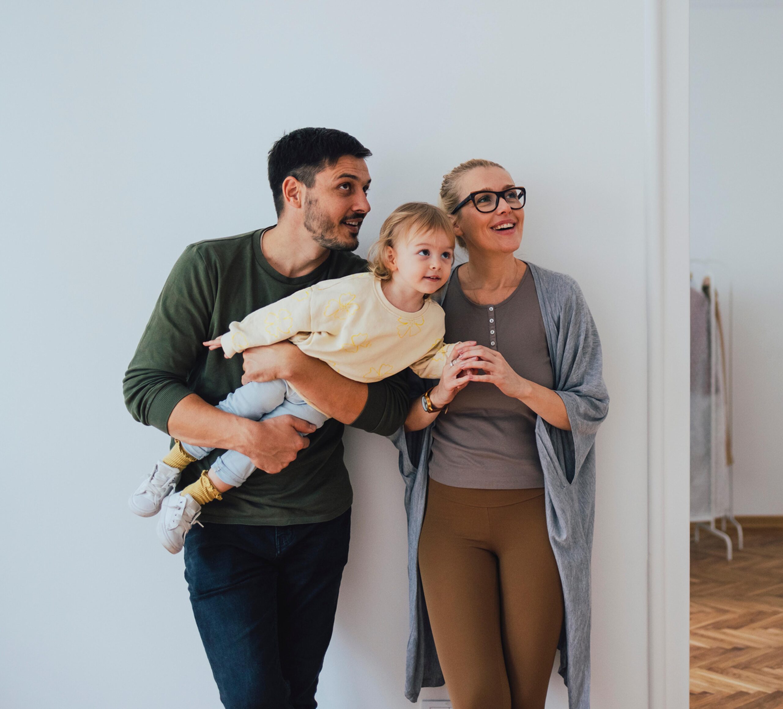 Ein junges Paar steht mit seinem kleinen Kind in einem modernen, hellen Zuhause. Der Mann mit dunklen Haaren und Bart trägt ein grünes Langarmshirt und hält das Kind spielerisch im Arm, während die Frau mit blonden Haaren, Brille und einem grauen Cardigan daneben steht und lächelnd in die Ferne schaut. Das Kind, in heller Kleidung mit gelben Socken, blickt neugierig in dieselbe Richtung. Die Szene strahlt Freude und Geborgenheit aus.